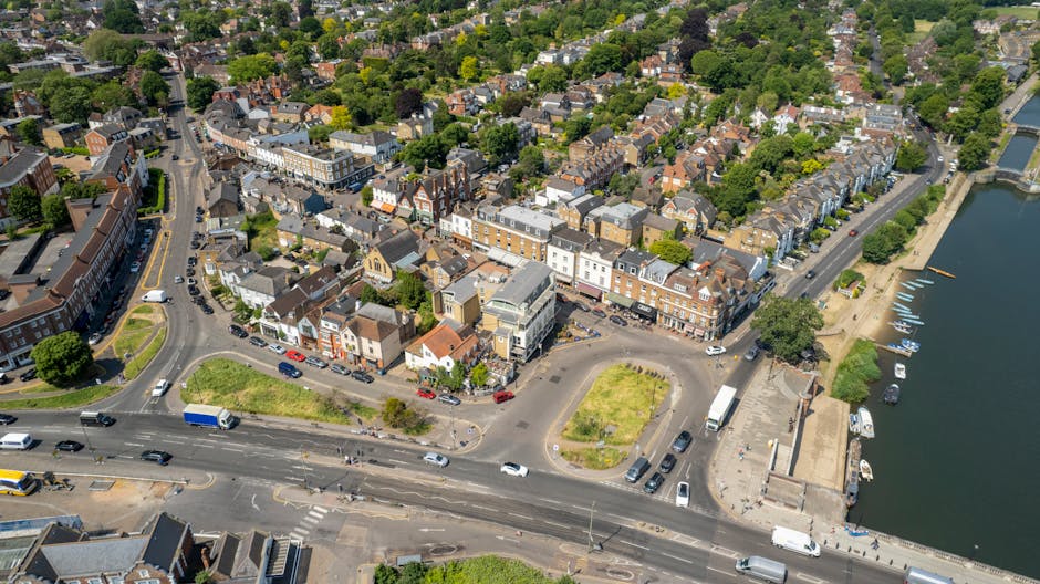 An aerial view of Colliers Wood High Street in London, showing a mix of terraced and commercial buildings along the street. The scene includes a small, landscaped roundabout with grass patches, vehicles parked along the roadside, and a river with boats moored along its bank to the right. The area is surrounded by lush green trees and residential houses, with a bright, clear sky overhead. This image visually highlights the urban environment of Colliers Wood, where professional cleaning services by Colliers Wood Carpet Cleaning may be needed for commercial and residential premises, particularly for surface cleaning and maintenance of carpets, floors, and internal spaces.