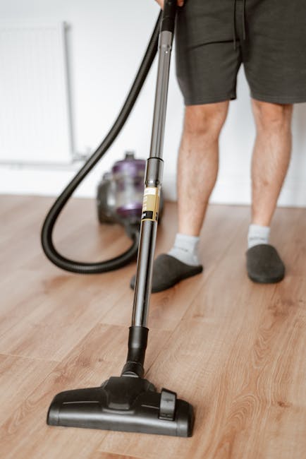 A person wearing gray shorts, a long sleeve shirt, and dark slippers is vacuuming a wooden floor in a room with white walls. The vacuum cleaner has a black floor head connected to a flexible hose and a metal tube, which the individual is holding to perform surface cleaning. A purple canister vacuum with a transparent dust container is visible in the background. The room appears tidy and well-lit, demonstrating professional cleaning processes suitable for residential environments. This image exemplifies deep cleaning and maintenance practices promoted by Colliers Wood Carpet Cleaning for high street shops and domestic spaces.
