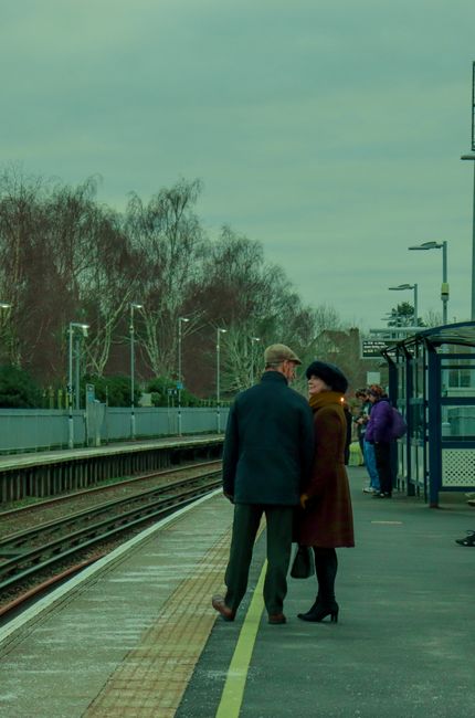 At Colliers Wood station, two commuters are engaged in conversation on the platform, which is clean and well-maintained with smooth, tiled surfaces. The platform features a yellow safety line along the edge and is equipped with modern lighting and a glass and metal shelter providing shelter for waiting passengers. In the background, additional passengers stand near the shelter, dressed in warm clothing suitable for cool weather. The tracks run parallel to the platform, and leafless trees and streetlights are visible beyond the fencing under an overcast sky. This setting exemplifies a tidy and orderly train station environment, reflecting effective maintenance and cleanliness supported by Colliers Wood Carpet Cleaning for local hygiene standards.