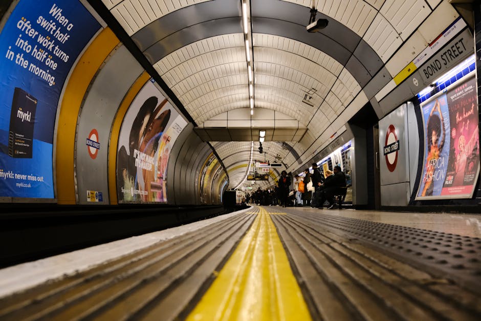 A spacious underground corridor at Colliers Wood station featuring polished light grey stone flooring with dark grey strip accents running parallel along the center. The walls are composed of smooth, reflective grey panels, with modern digital advertising screens mounted at intervals. The ceiling is painted black, with sleek, linear lighting fixtures providing bright illumination across the space. At the end of the corridor, there are two lifts flanked by emergency exit doors and signage indicating platform directions. The area appears clean, well-maintained, with no visible dust or debris, consistent with professional surface cleaning and sanitisation by Colliers Wood Carpet Cleaning to ensure it remains safe and hygienic for commuters.