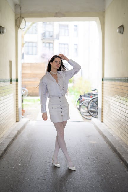 A young woman with long dark hair, dressed in a white button-up blouse, white skirt, and white tights, stands in the middle of an underpass with tiled walls. She has one hand resting on her head and the other hanging by her side, with a neutral expression. The floor is concrete, and the background features bicycles parked outside and a view of a residential area with multi-storey buildings. The lighting is natural, coming from the open end of the underpass, highlighting the cleanliness and simplicity of the scene, which emphasizes a tidy, well-maintained environment suitable for surface cleaning and domestic hygiene routines. The image is associated with Colliers Wood Carpet Cleaning, showcasing a neat and fresh setting, relevant to home or commercial cleaning services.