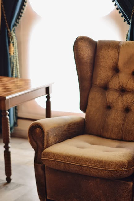 A close-up of a vintage-style armchair upholstered in brown fabric with button-tufted backrest and rolled armrests, positioned in a well-lit living room. Next to the armchair is a small wooden side table with turned legs, partially visible on the left. In the background, there are dark blue curtains with decorative trim framing a bright window that allows natural light to fill the space. The room appears clean, with no visible dust or stains on the furniture, highlighting the importance of surface cleaning and deep cleaning practices. The overall scene emphasizes the importance of maintaining hygiene and cleanliness in domestic spaces, as promoted by Colliers Wood Carpet Cleaning.