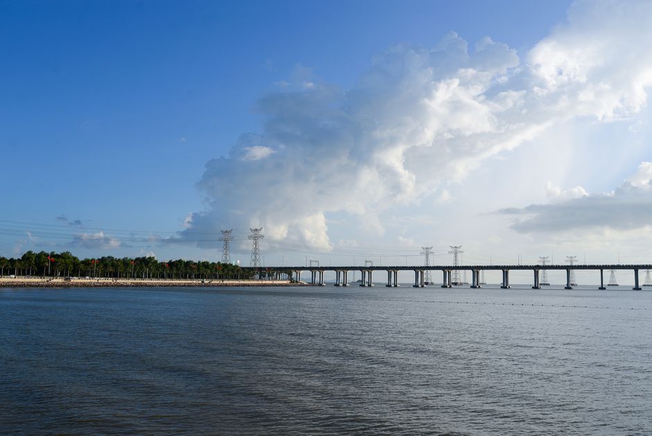A wide view of a calm water body with a distant bridge supported by multiple concrete pillars stretching across the water. The shoreline features a line of green trees, and the sky overhead is partly cloudy with large, white, billowing clouds and patches of blue. Bright sunlight illuminates the scene, highlighting the clear, smooth surface of the water, which reflects the sky and clouds. This landscape illustrates a peaceful outdoor setting, with the bridge and power line pylons in the background emphasizing infrastructure. Colliers Wood Carpet Cleaning would use such an image to promote surface cleaning and maintenance services in outdoor or associated indoor environments, aligning with deep cleaning and sanitisation themes.