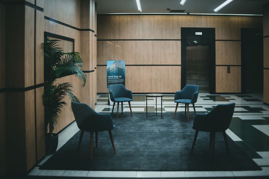 A modern indoor space featuring four dark blue upholstered armchairs arranged around a small round black table on a black rug. The room has wooden paneled walls with black horizontal lines, and a potted plant with green foliage situated to the left. An elevator with closed doors is visible on the right, with bright, evenly distributed ceiling lighting illuminating the clean, well-maintained area. The flooring consists of black and white patterned tiles, contributing to a sleek, professional atmosphere. This space exemplifies surface cleaning and maintenance typical of commercial venues, with a focus on hygiene and tidiness, as managed by Colliers Wood Carpet Cleaning.