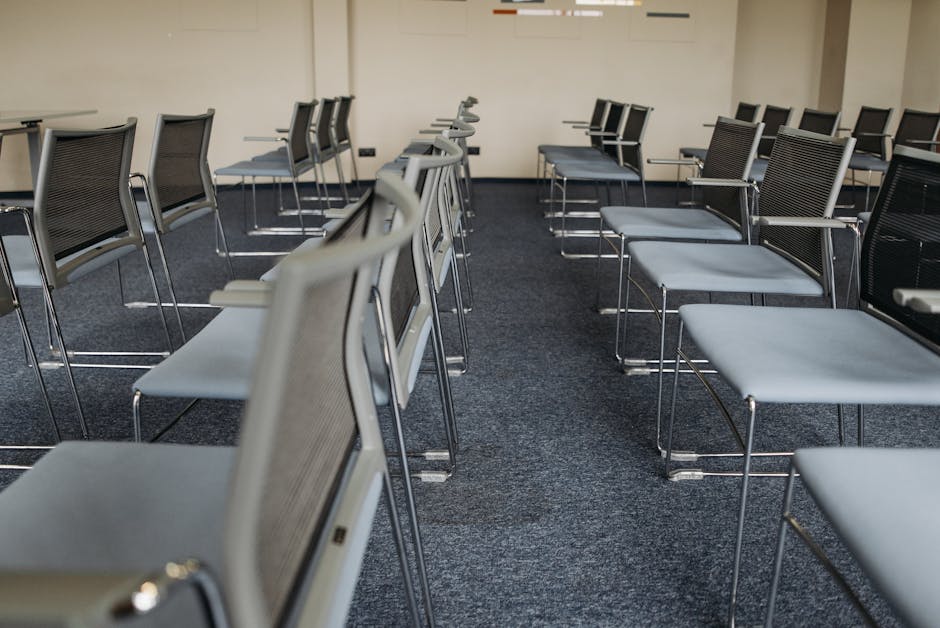 Image depicting a neatly arranged conference or event room with multiple rows of grey chairs featuring metal frames and mesh backs, aligned on a dark grey carpeted floor. The chairs are evenly spaced, and the room is illuminated with soft, natural light. The plain beige or off-white walls in the background provide a neutral setting, emphasizing the cleanliness and orderly layout. The space appears well-maintained and free of dust or clutter, illustrating professional surface cleaning and hygiene standards. Colliers Wood Carpet Cleaning specializes in deep cleaning and sanitisation services for venues such as this, ensuring hygienic, spotless conditions for events at locations like Plough Lane. The clean and fresh environment highlights the company’s proficiency in domestic and commercial cleaning.