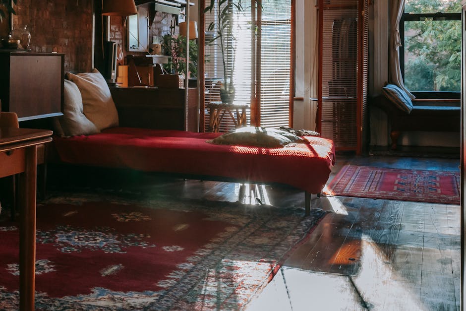 A cozy living room with a wooden floor, partially covered by patterned area rugs in red and blue tones. A red sofa with white cushions is positioned against a brick wall with shelves and decorative items. Sunlight filters through wooden blinds on large windows, casting natural light and shadows across the space. A side table and a wooden bench with a cushion are visible near the window. The overall scene reflects a clean and well-maintained domestic setting, showcasing surface cleaning and deep cleaning practices as part of routine garden or home sanitisation, with attention to maintaining hygiene and visual appeal. Colliers Wood Carpet Cleaning’s expertise ensures the surfaces are free of dust and dirt, promoting a healthy living environment.
